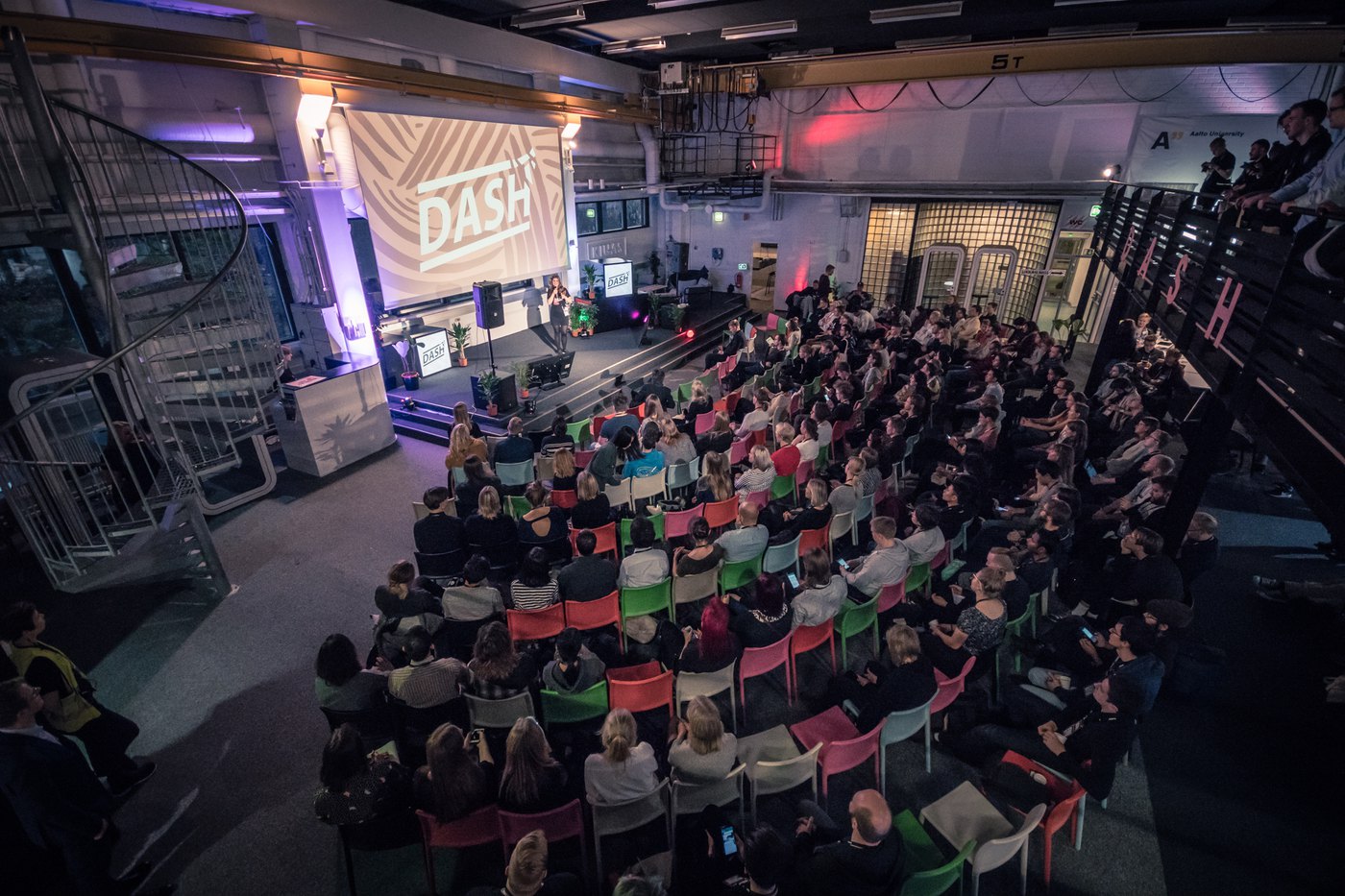 The Dash main hall during a talk: a speaker on a low stage in front of a large projected Dash logo, with a full house of several hundred people seated in rows of coloured chairs, more watching from a balcony, and an Aalto University banner on the back wall