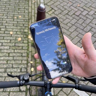 A phone held above bicycle handlebars on a wet brick street, its screen showing an app with a bicycle illustration and the name Speedy McNeedy